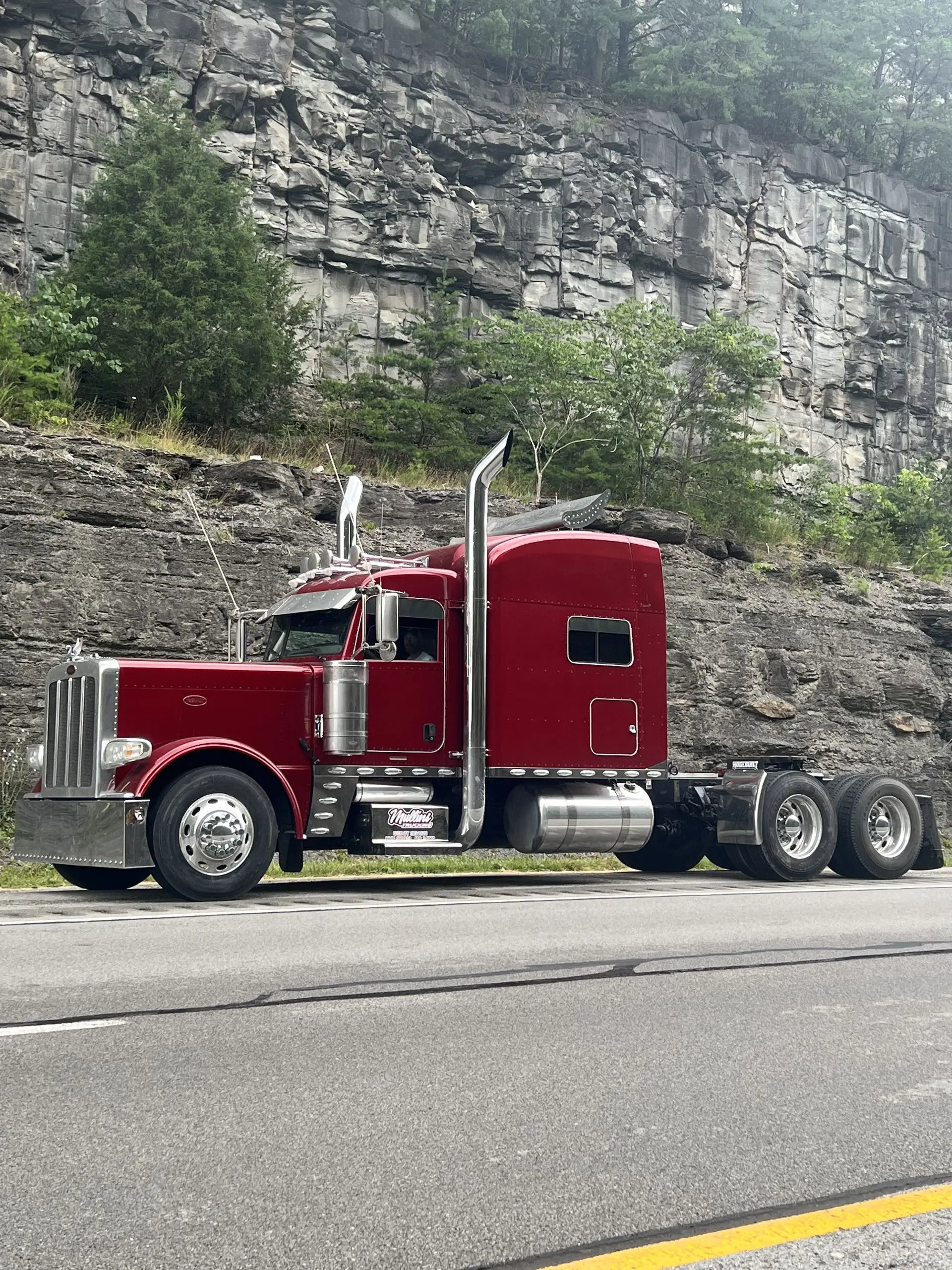 Maroon Mullins Trucking Peterbilt bobtail on a Kentucky highway shoulder below a rock cut
